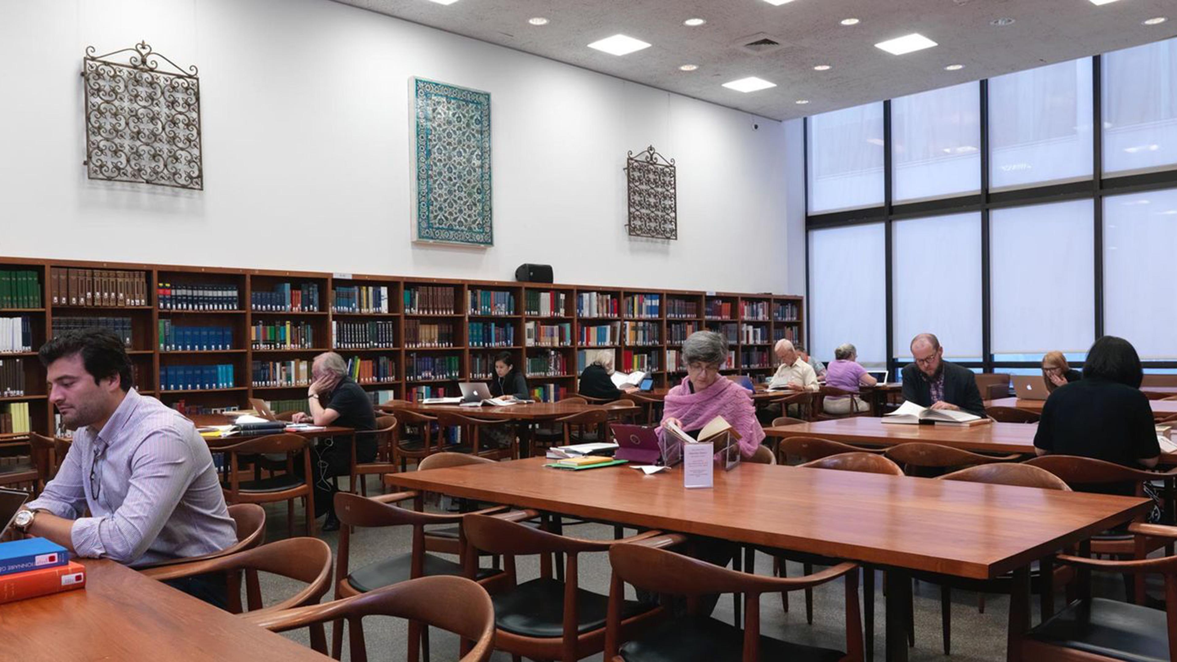 Individuals seated at large wooden desks reading with a wall of bookshelves and windows in the background. 