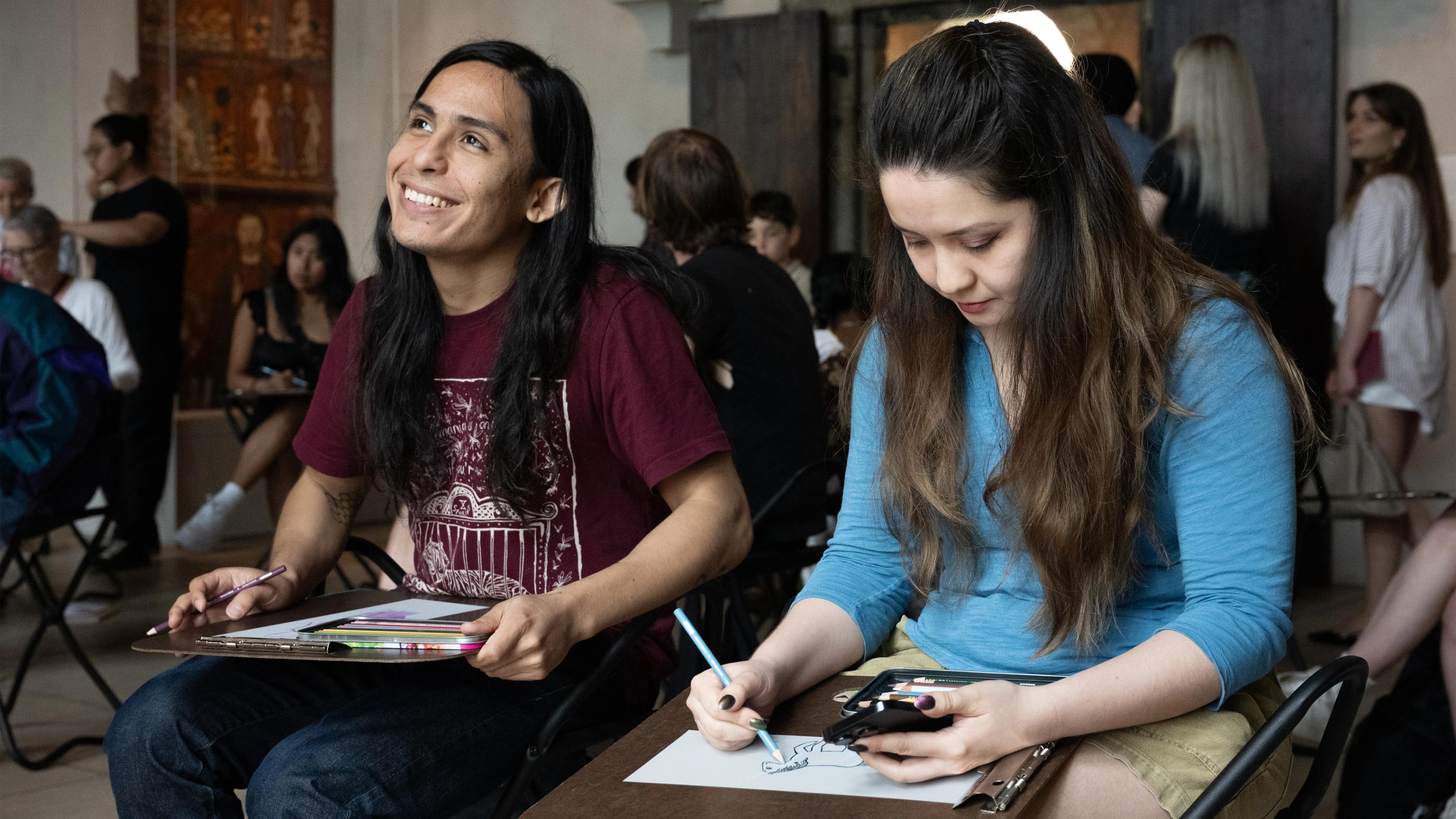 Two young people engaged in an art class, one drawing with a pencil and the other holding their work and looking up smiling, amid a group of peers.