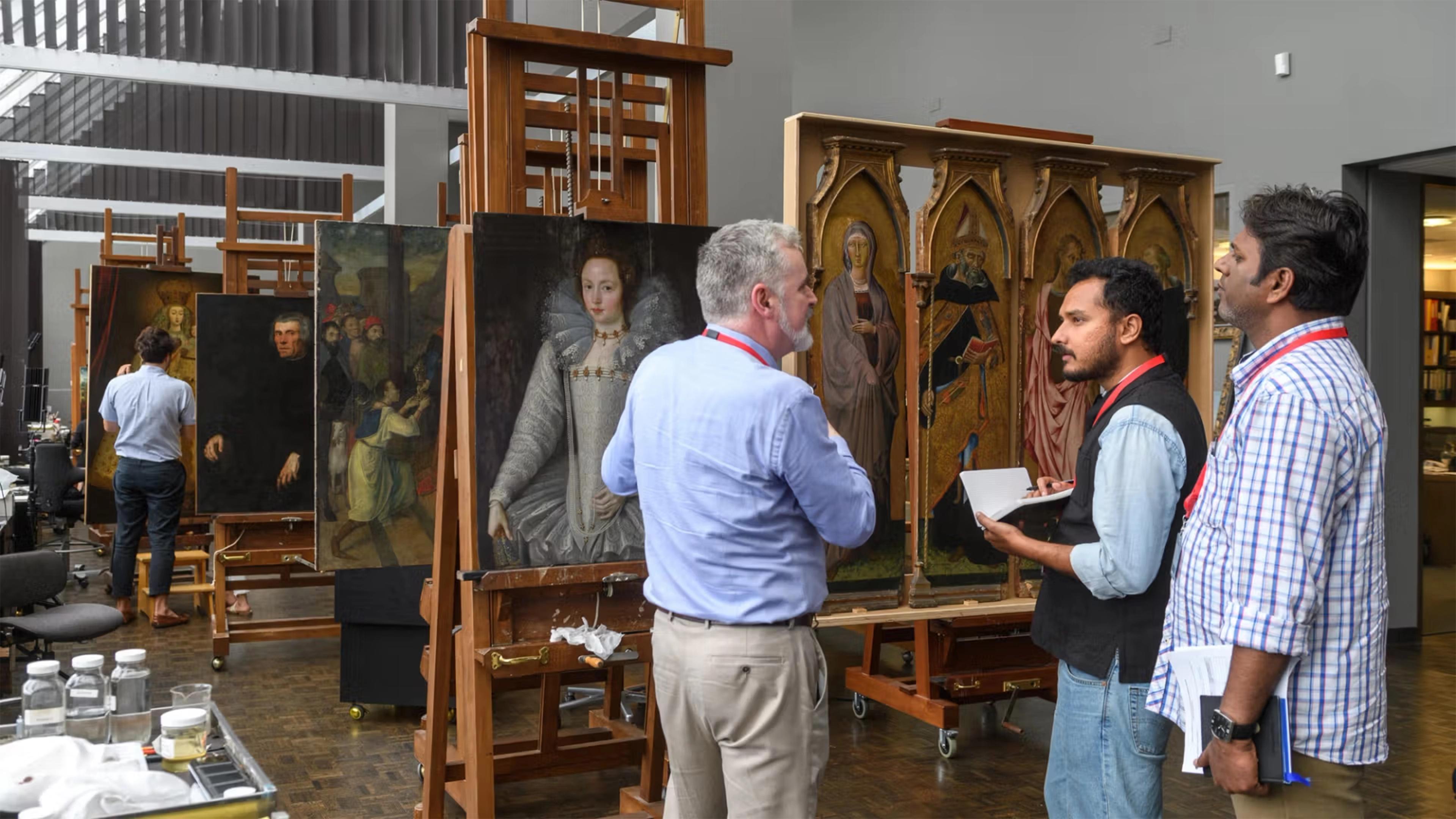 Three men stand in a painting restoration workshop, examining large paintings on easels. One man in the background is in a light blue shirt and khaki pants, and is seen from behind. The other two face the artwork; one with a notebook, the other listening. They are surrounded by framed portraits paintings and altarpieces.