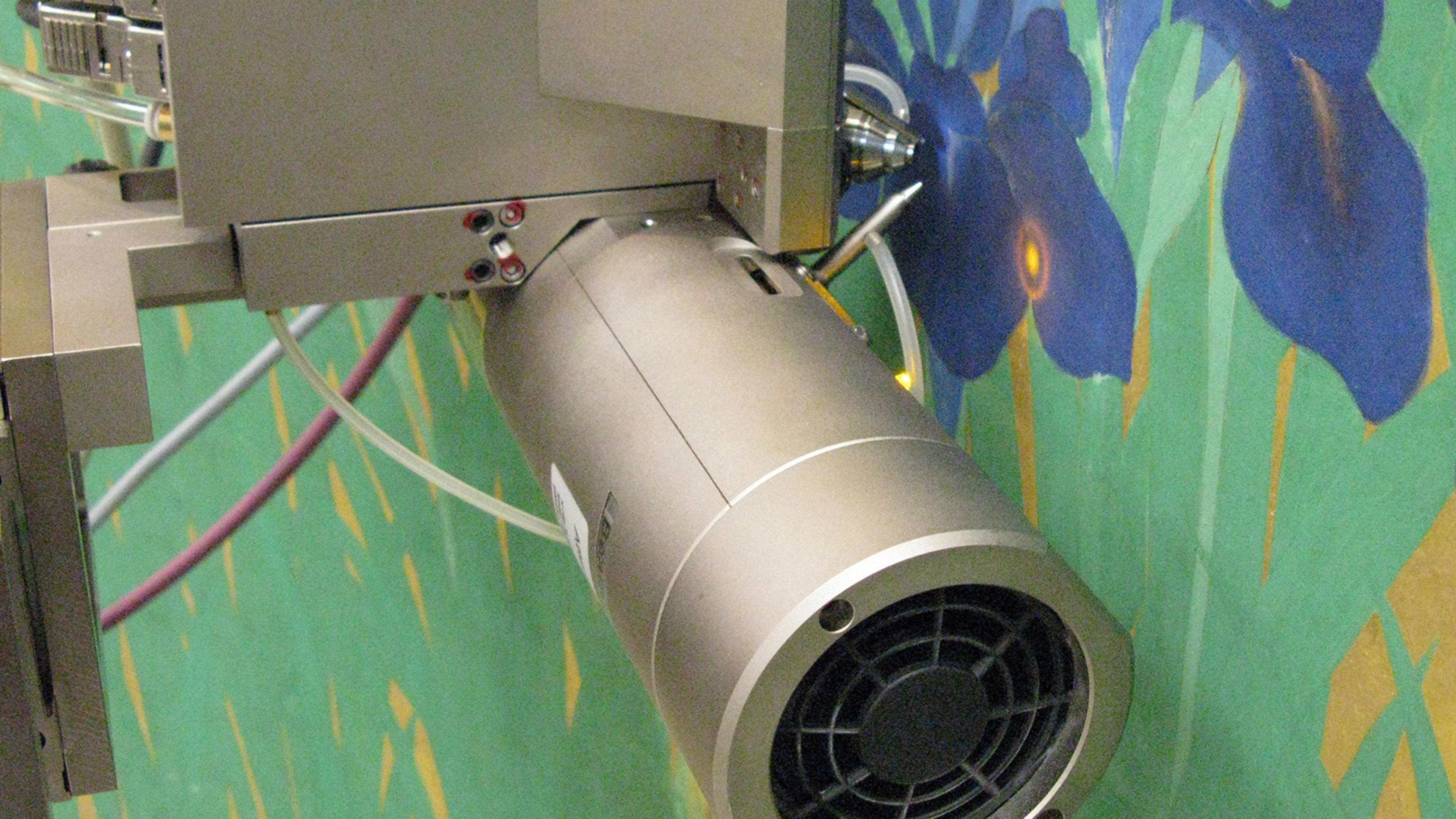 Photographed up-close, from three-quarter view, a piece of silver scientific equipment shines a precise bulls-eye light on a painting of blue irises.