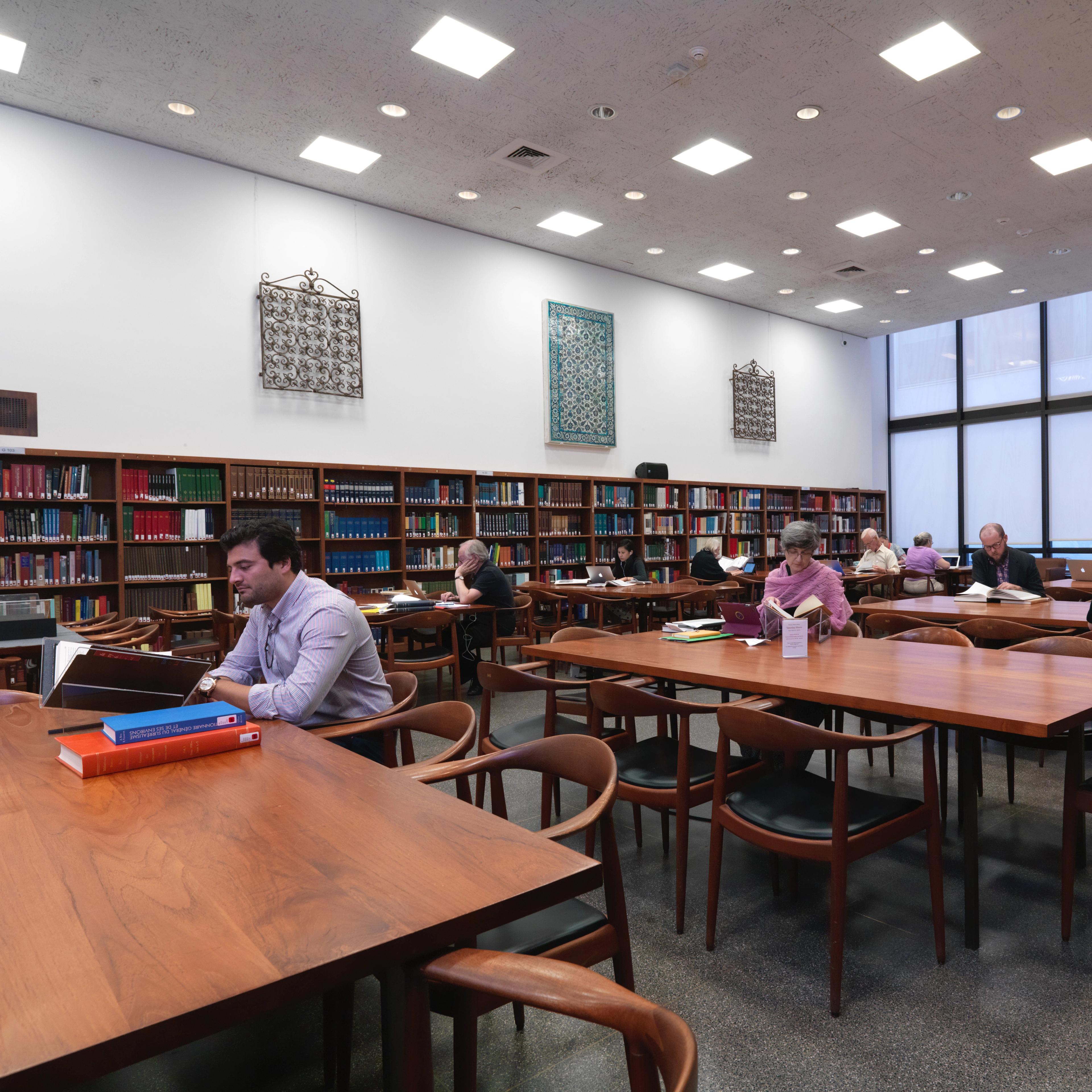 A spacious library with wooden tables, shelves filled with books along the back wall, and floor-to-ceiling windows on the right wall. Several individuals are reading books at the tables.