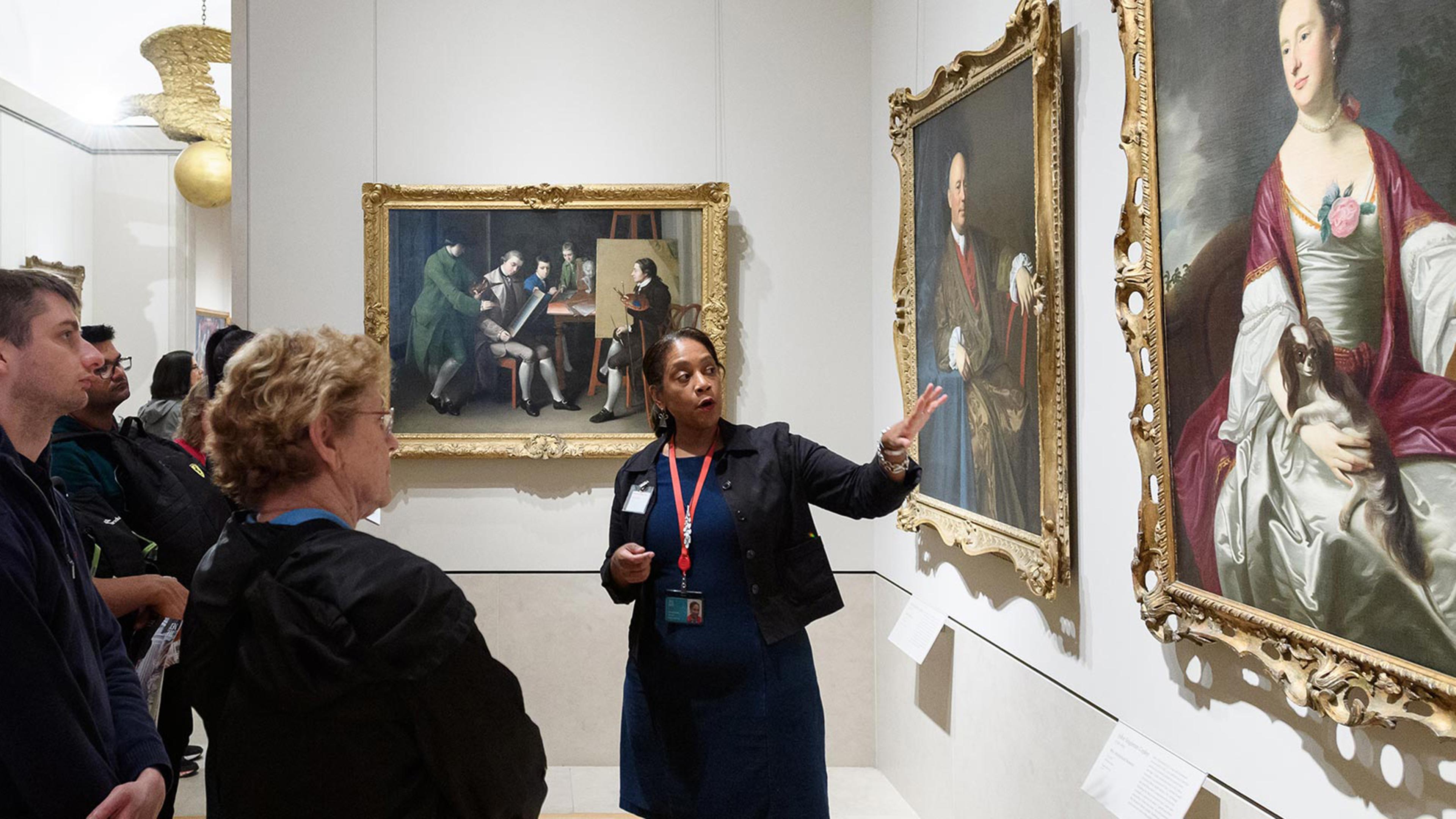 A volunteer leads a group of visitors on a tour in the American Wing. The volunteer is gesturing towards and discussing an oil painting in The Met collection.
