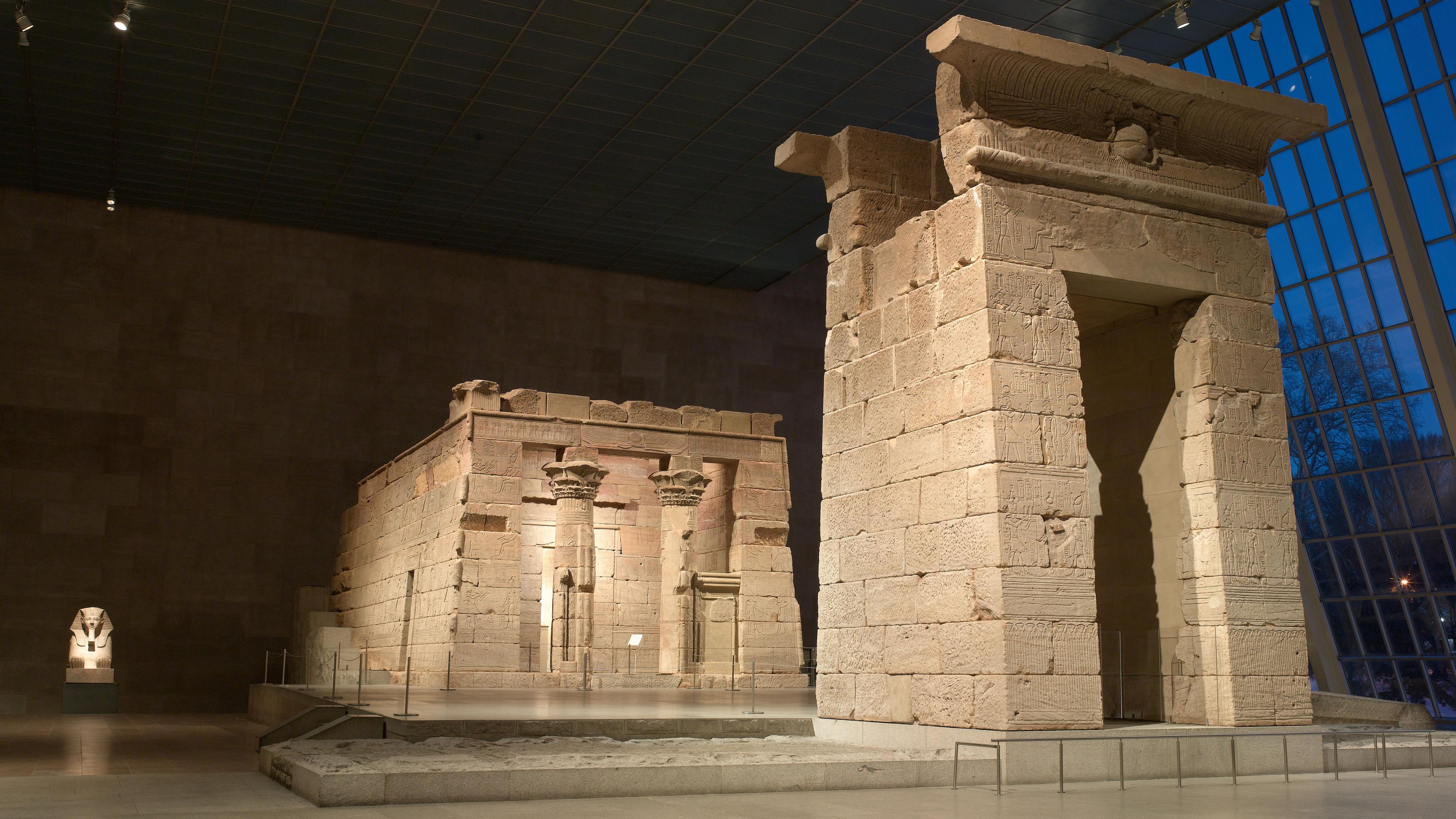 The Temple of Dendur, an ancient Egyptian sandstone temple, displayed at The Metropolitan Museum of Art. Dramatic lighting highlights its intricate carvings and grand gateway, contrasting with the dark blue evening sky visible through the glass wall behind it.