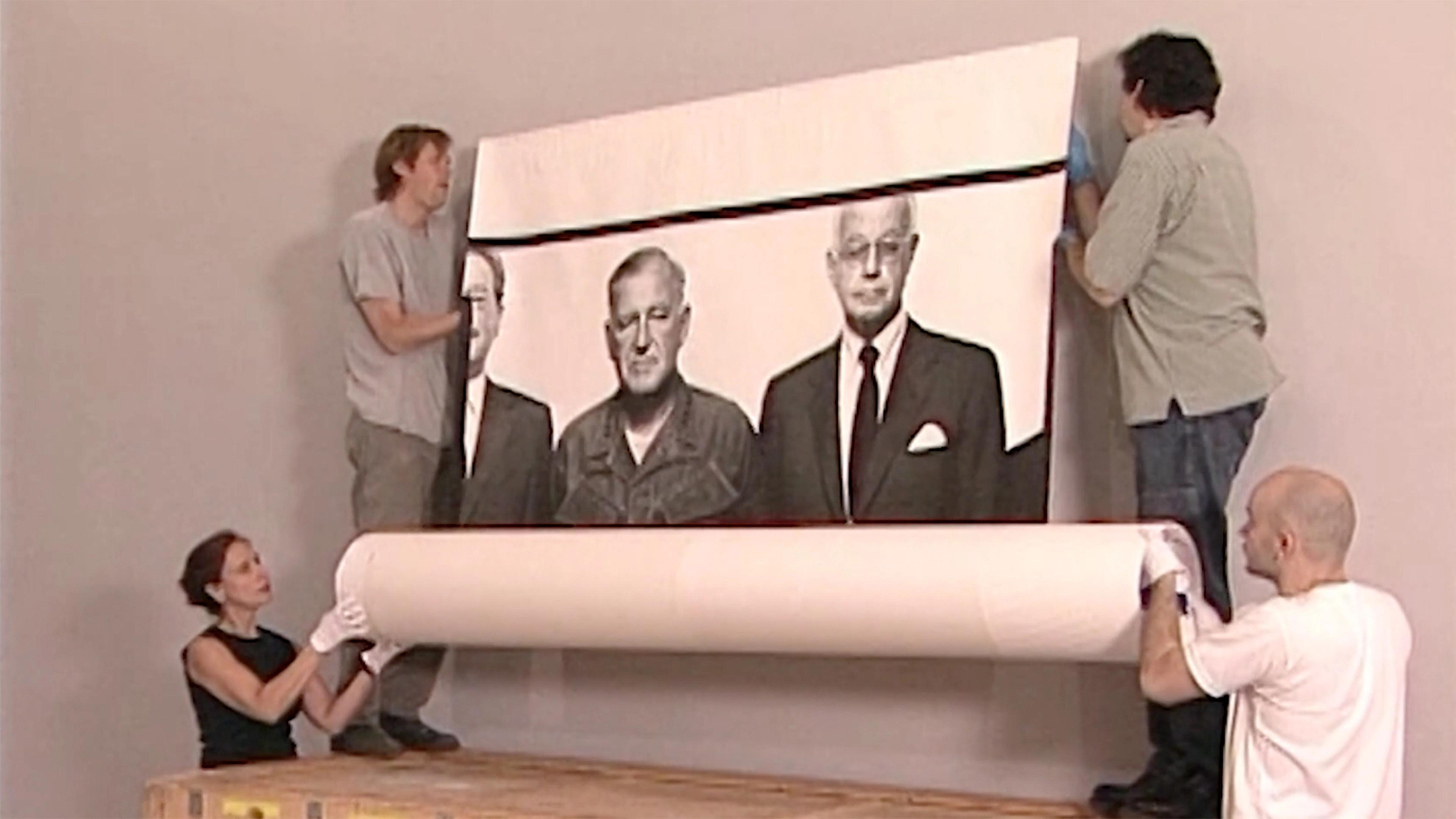Two people are standing on a crate to attach a large, black-and-white print to the wall, while two stand below holding the rolled part of the print.