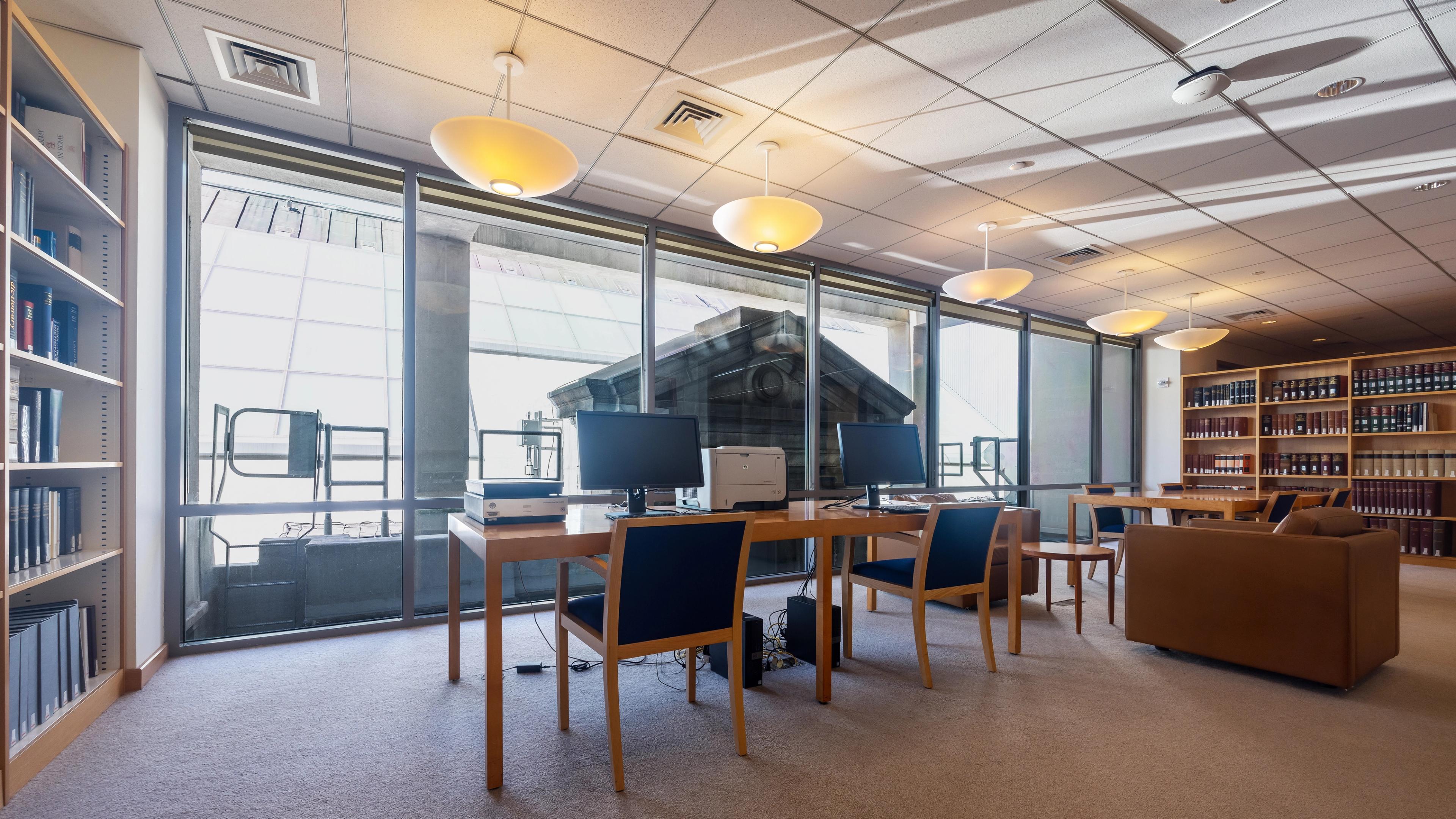 An open study space with computers and printers on long tables, cozy seating, and large windows providing natural light and a city view. Shelves filled with books line the far left and far right walls.