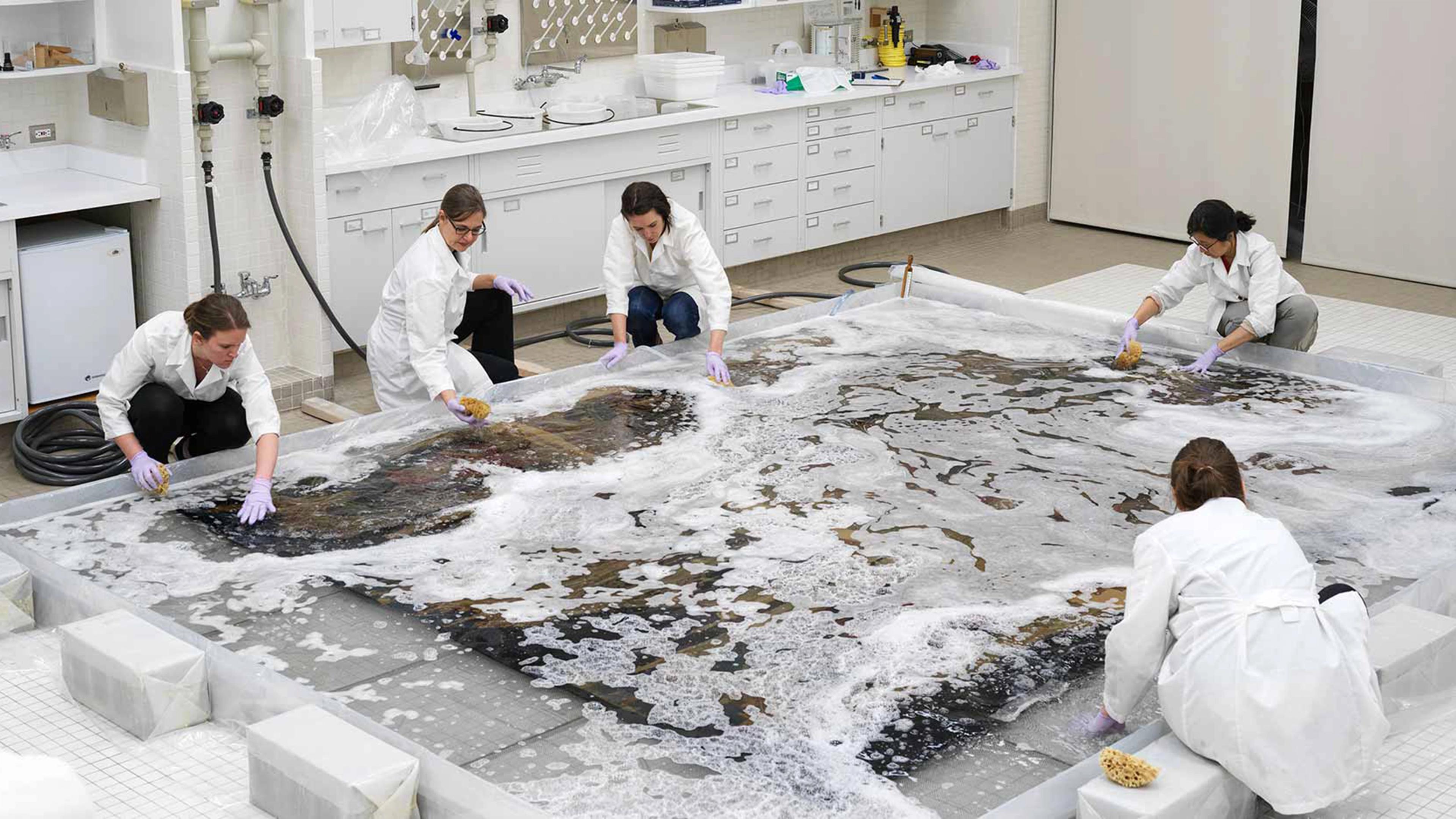 5 individuals in white lab coats kneel over a large, flat textile to clean it with foam and water, using sponges and other tools.