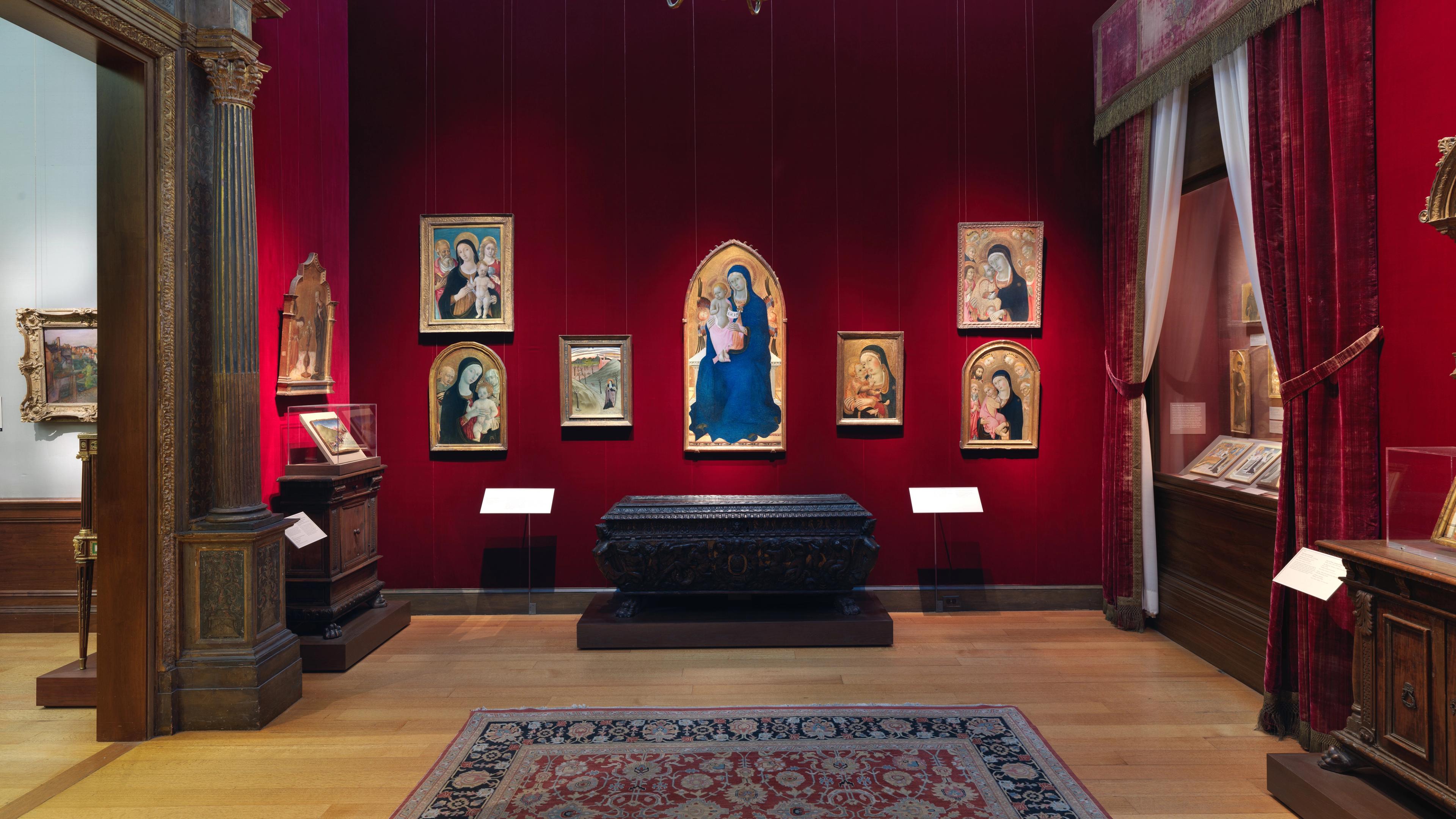 A room within the Robert Lehman Collection at The Met with red walls displays framed Renaissance religious paintings, a dark ornate chest, and wooden furniture.