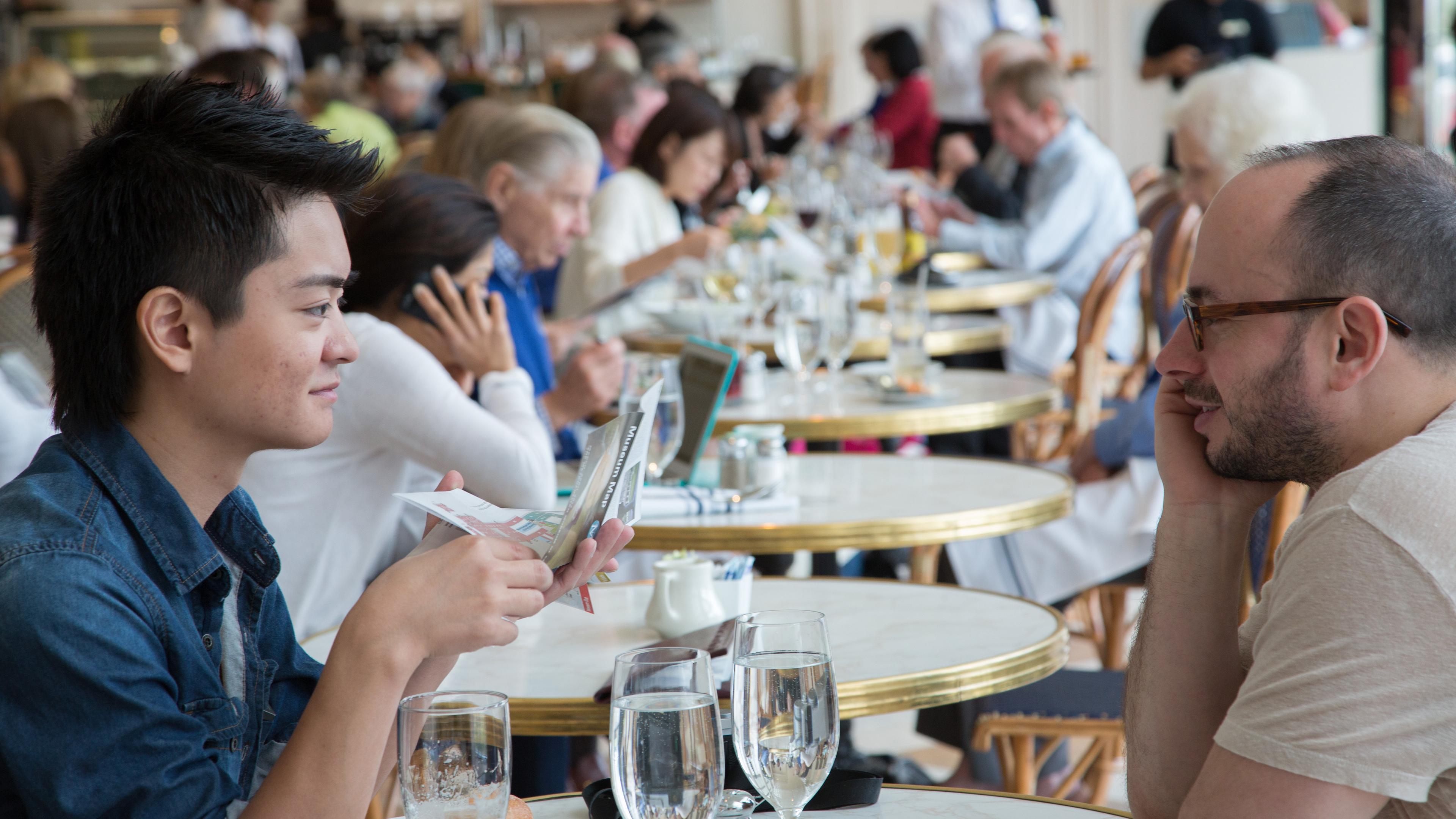 Two Museum visitors sit at a table in the American Wing Cafe in The Met, talking over drinks.